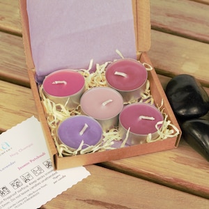 May include: A set of five tealight candles in a cardboard box with a lavender, hay, jasmine, and patchouli scent. The candles are in a variety of colours, including pink, purple, and lavender. The box is sitting on a wooden surface.