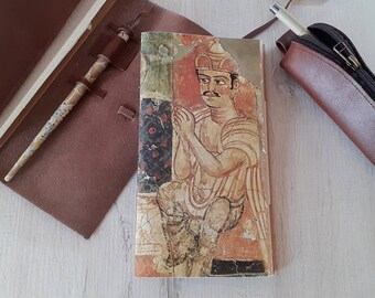 Peut inclure: Un carnet vintage avec une couverture en cuir marron et une illustration colorée d'un homme portant une couronne. L'illustration est peinte dans des tons de rouge, jaune et bleu.