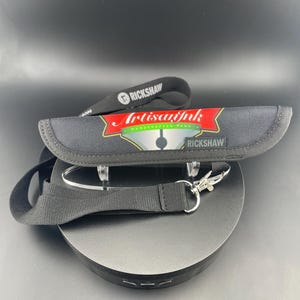 Puede incluir: Estuche negro para bolígrafos con el texto "Artisan Ink Handcrafted Pens" y la marca "Rickshaw". El estuche está sobre un soporte negro con un cordón negro y un cierre plateado.