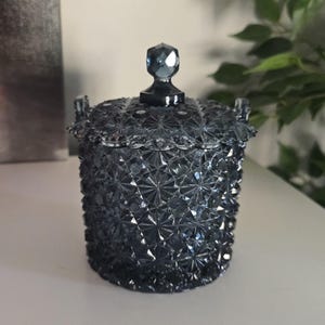 Puede incluir: Un tarro azul oscuro de estilo vintage con tapa. El tarro tiene un patrón texturizado de estrella y asas pequeñas. La tapa está rematada con un pomo facetado en forma de gema. El tarro es probablemente de vidrio o cristal.