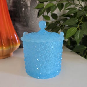 Puede incluir: Un tarro azul claro con tapa, con un diseño de estrella texturizado. El tarro tiene un pequeño asa en la tapa y dos asas laterales pequeñas. El tarro es probablemente vintage.