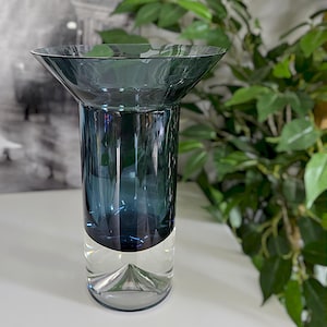 May include: A tall, elegant glass vase with a flared top and a cylindrical body. The upper portion is a deep teal color, transitioning to clear glass at the base. The vase is designed for floral arrangements or decorative display.