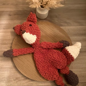 May include: A crocheted fox plush toy with a red body, white face, and brown accents. The toy is lying on a wooden surface.