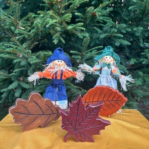 May include: Two scarecrow dolls with straw hair and painted faces stand on a yellow surface, with wooden leaf cutouts in front. The scarecrows wear blue and orange outfits. The background is a green evergreen tree.