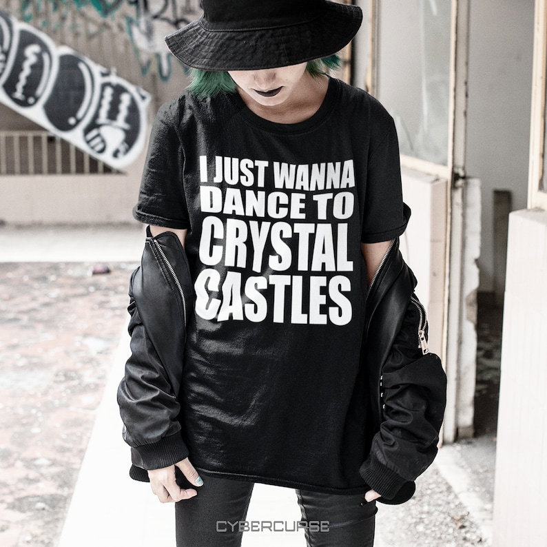 May include: A black t-shirt with white text that reads "I just wanna dance to crystal castles". The person wearing the shirt is wearing a black hat and black pants.