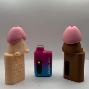 Puede incluir: Se muestran tres artículos novedosos sobre un fondo blanco. Dos son objetos fálicos beige y marrones con tapas rosas. El tercero es un pequeño dispositivo de vapeo rectangular con un degradado de azul y rosa, con la palabra "DABBAR" impresa.