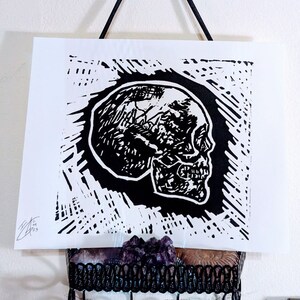 May include: Black and white linocut print of a skull in profile. The skull is detailed and has a textured look. The print is on white paper and has a black border.
