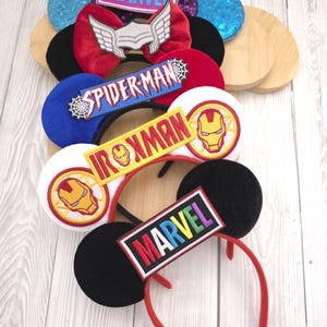 Pode incluir: Uma coleção de tiaras com tema Marvel com orelhas do Mickey Mouse. As tiaras apresentam designs de personagens como Homem-Aranha, Homem de Ferro e Thor, com o logotipo da Marvel. As orelhas são pretas, azuis e vermelhas, algumas enfeitadas com lantejoulas.