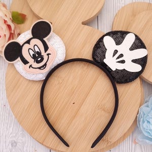 Può includere: Una fascia nera con due decorazioni a orecchie di Topolino. Una presenta il volto di Topolino su uno sfondo di paillettes bianche, l'altra una mano bianca su uno sfondo di paillettes nere. La fascia è su un ritaglio di legno a forma di Topolino.