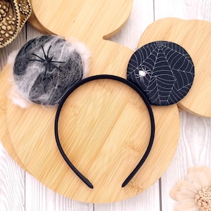 May include: A black headband with two circular decorations. One has a spiderweb design with a small white spider, the other a black spider and white web. A Halloween-themed accessory.
