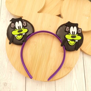 May include: A purple headband featuring two black cartoon character ears with green and yellow accents. The ears are attached to a purple plastic headband. The headband is resting on a wooden surface.