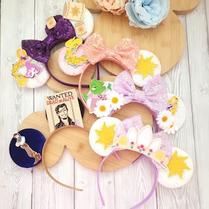May include: A collection of themed headbands with decorative bows, ears, and embellishments. The headbands feature characters and symbols from a popular animated film. The color palette includes pink, purple, yellow, and white, with accents of glitter and sequins. A small sign reads "WANTED DEAD OR ALIVE."