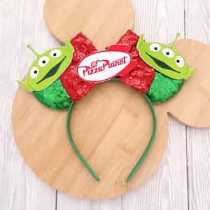 May include: A green headband featuring two alien figures and a red sequined bow. The bow has a white oval with the words "Pizza Planet" in red. The headband is green and the aliens have three eyes.