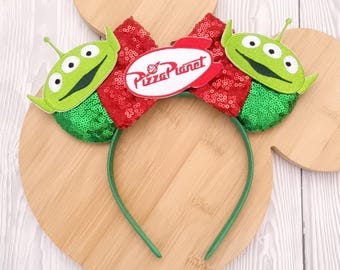 Puede incluir: Una diadema verde con dos figuras de extraterrestres y un lazo rojo con lentejuelas. El lazo presenta un óvalo blanco con las palabras "Pizza Planet" en rojo. La diadema es verde y los extraterrestres tienen tres ojos.
