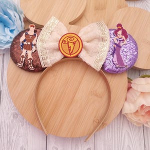 May include: A pair of light brown mouse ears with a white bow and gold trim. The bow has a gold lightning bolt emblem in the center. One ear features a purple sequined design with a cartoon image of a woman in a purple dress. The other ear features a brown sequined design with a cartoon image of a man in a brown tunic and sandals.