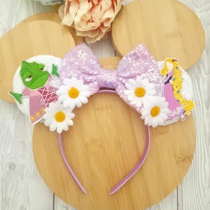 May include: A lavender sequined bow headband with white daisy accents and character details. The headband features a light purple band and white ear details with a frog and a princess. The bow is decorated with sequins.