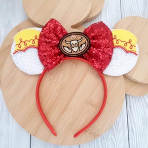May include: A red sequined bow headband with white sequined ears and yellow accents. The centre of the bow features a brown oval with a bull's head design. The headband is red and sits on a wooden surface.