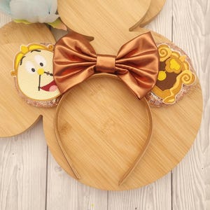 May include: A gold headband with attached ears featuring embroidered character designs. A large, copper-colored satin bow sits at the center. The ears have a sequined border. The background is a light wood grain.