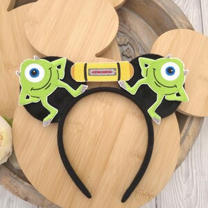 May include: A black headband featuring two circular ears decorated with embroidered green one-eyed monster patches. Between the ears is a yellow rectangular patch with a central graphic design. The headband has a thin, flexible black frame.