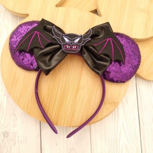 May include: A purple headband with sequined ears and a black satin bow. The bow features a bat-like embroidered design with purple accents. The headband is a solid purple color, creating a themed accessory.
