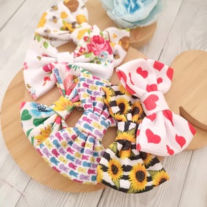 May include: Several fabric hair bows with diverse patterns, such as floral, sunflowers, and hearts. The bows are displayed on a wooden surface, with a light blue flower in the background. The bows are in various colours.