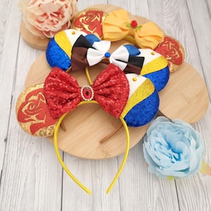 Puede incluir: Un conjunto de tres pares de orejas de ratón con diferentes diseños. Las orejas están hechas de tela y lentejuelas y presentan una combinación de colores rojo, amarillo, azul y blanco. Cada par tiene un lazo diferente, incluyendo un lazo rojo, un lazo amarillo y un lazo blanco con detalles azules. Las orejas están unidas a una diadema amarilla.