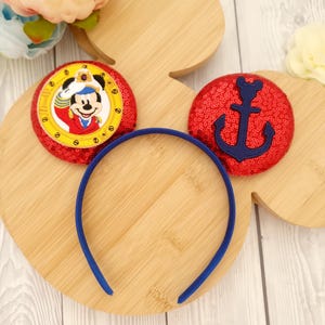 Pode incluir: Uma tiara azul com dois enfeites de orelhas do Mickey Mouse vermelhas com lantejoulas. Uma orelha apresenta o Mickey Mouse com uma roupa de marinheiro, e a outra tem um desenho de âncora azul marinho. As orelhas estão em uma base de madeira em forma de Mickey Mouse.