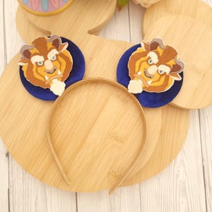 Könnte beinhalten: Ein beiges Stirnband mit zwei Plüschohren des Biest-Charakters. Jedes Ohr ist ein braunes und gelbes Biest-Gesicht auf einer dunkelblauen kreisförmigen Basis. Das Stirnband und die Ohren befinden sich auf einer hellbraunen Holzoberfläche.