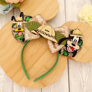 Può includere: Una fascia per capelli stravagante con un grande fiocco di paillettes dorate e verdi, ornata da immagini di personaggi dei cartoni animati e cappelli da safari. La fascia ha una fascia verde ed è progettata per un look giocoso.