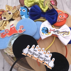 May include: A collection of character-themed headbands featuring various designs including a green sequin bow with a sea turtle, a blue velvet bow, an orange fish, a white bow with the text Just keep swimming, and black ears with seagulls and the text MINE!