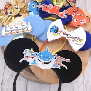 Op de afbeelding: Een collectie haarbanden met karakterthema's en diverse aquatische ontwerpen. De haarbanden bevatten elementen zoals lovertjesstrikken, pluche oren en geborduurde patches van haaien, meeuwen met de tekst Mine en vissen met de tekst Just Keep Swimming.