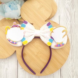 May include: A purple headband featuring a white sequined bow and decorative cutouts of a teapot and teacup. The teapot and teacup are white with gold, purple, yellow, and blue accents. The headband is on a wooden surface.