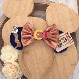 May include: A headband featuring two circular ears with a metallic copper bow in the centre. The left ear displays an embroidered character in a blue vest and tan trousers. The right ear features a rectangular tag with the text WANTED DEAD or ALIVE.