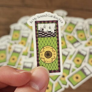 May include: A sticker featuring a green and purple design resembling a music player. The sticker has the text "In My Sound Garden Era" at the top. The music player design has a circular button with the word "MENU" at the bottom.