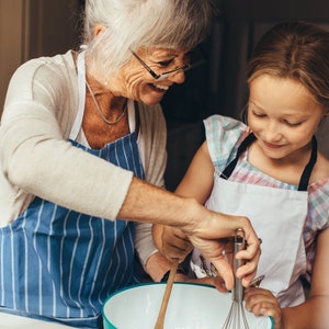 May include: An older woman and a young girl wearing aprons are baking together. The woman is holding a whisk and the girl is holding a wooden spoon. They are both smiling and looking at each other.
