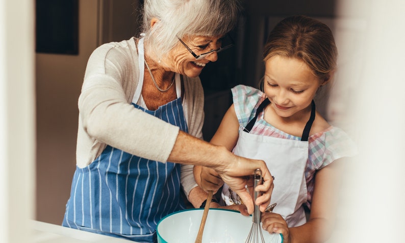 May include: An older woman and a young girl wearing aprons are baking together. The woman is holding a whisk and the girl is holding a wooden spoon. They are both smiling and looking at each other.