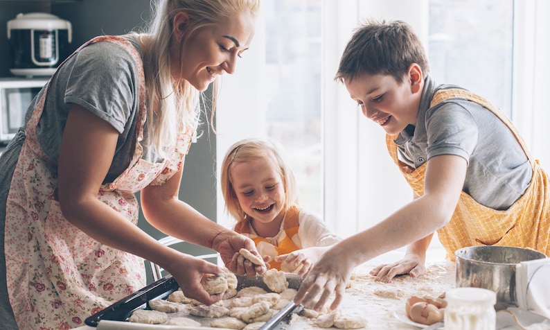 May include: A family of three, a woman and two children, are in a kitchen preparing food. They are all wearing aprons and smiling. The woman is holding a piece of dough, while the children are helping to shape it. There is a baking sheet with dough on it in the foreground.