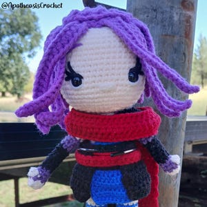 May include: A handmade crochet doll with purple hair, black eyelashes, and a red scarf. The doll wears a black jacket, blue trousers with white accents, and black boots. The doll is standing on a black base. The text "@ApotheosisCrochet" is visible.