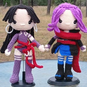 May include: Two handmade crochet dolls. One doll has black hair, white hoop earrings, and a purple outfit with a red belt. The other doll has purple hair, a red scarf, and a blue and black outfit. Both dolls are on black stands.