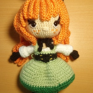 May include: A crocheted doll with orange hair, black cat ears, a green dress, and a black belt. The doll has black eyes and a white face.