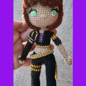 May include: Crocheted doll with brown hair and green eyes. The doll wears a black outfit with gold and purple accents. The doll's hair is styled in a curly bob. The doll is handmade and has a whimsical appearance.