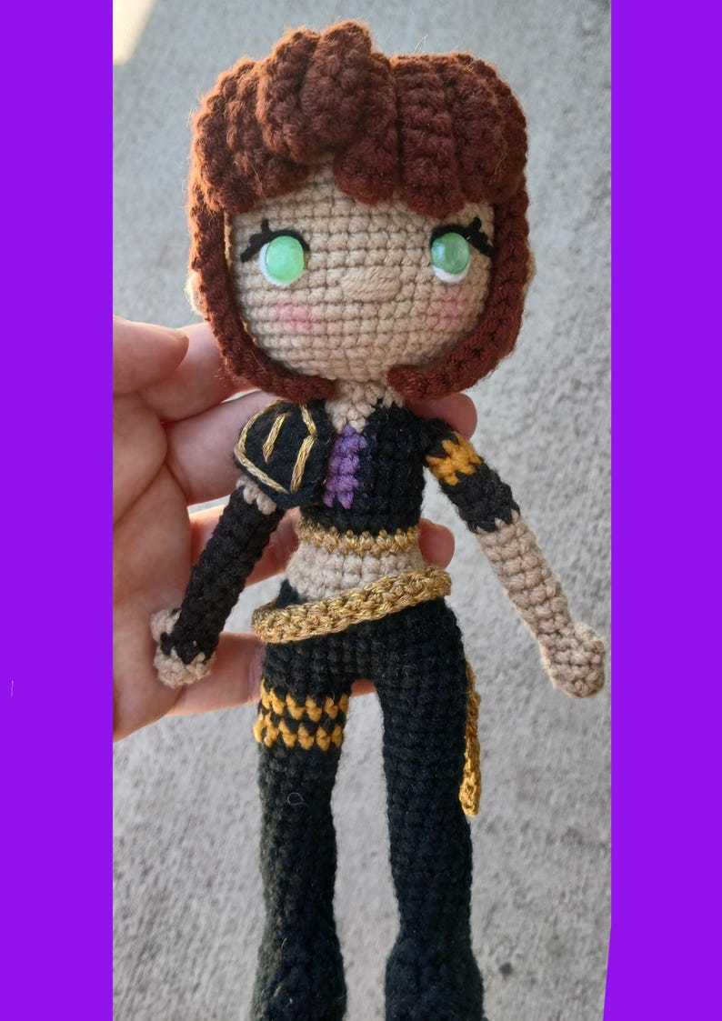 May include: Crocheted doll with brown hair and green eyes. The doll wears a black outfit with gold and purple accents. The doll's hair is styled in a curly bob. The doll is handmade and has a whimsical appearance.