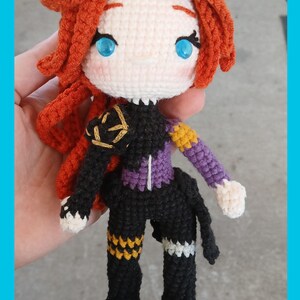 May include: Crocheted doll with red hair, blue eyes, and a black and purple outfit. The doll has a white face and hands, with gold and yellow accents on the clothing. The doll is handmade and appears to be a character from a video game or anime.