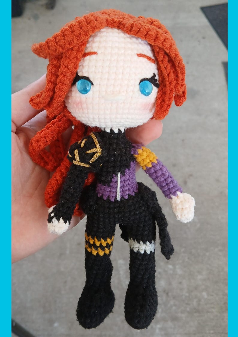May include: Crocheted doll with red hair, blue eyes, and a black and purple outfit. The doll has a white face and hands, with gold and yellow accents on the clothing. The doll is handmade and appears to be a character from a video game or anime.