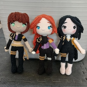 May include: Three handmade crochet dolls with detailed outfits. Each doll has a unique hairstyle and eye color: brown, red, and black hair with green, blue, and black eyes, respectively. The dolls wear black and gold outfits with intricate detailing.