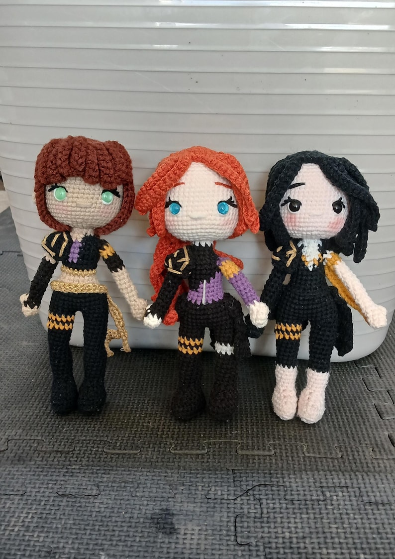May include: Three handmade crochet dolls with detailed outfits. Each doll has a unique hairstyle and eye color: brown, red, and black hair with green, blue, and black eyes, respectively. The dolls wear black and gold outfits with intricate detailing.