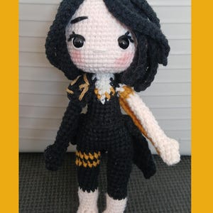 May include: A handmade crochet doll with black hair and a black outfit with gold accents. The doll has a light skin tone, rosy cheeks, and large eyes. The doll's arms and legs are a light beige color. The doll is standing upright.