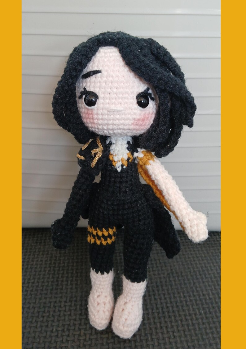 May include: A handmade crochet doll with black hair and a black outfit with gold accents. The doll has a light skin tone, rosy cheeks, and large eyes. The doll's arms and legs are a light beige color. The doll is standing upright.