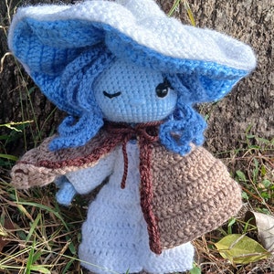 May include: A crocheted doll wearing a blue and white hat, a brown and white cape, and a white dress. The doll has blue hair and is winking.