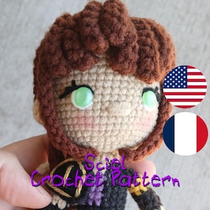 May include: A handmade crochet doll with brown hair, green eyes, and rosy cheeks. The doll is wearing a black and purple outfit. The text "Sciel Crochet Pattern" is visible on the image. The doll is held in a person's hand.